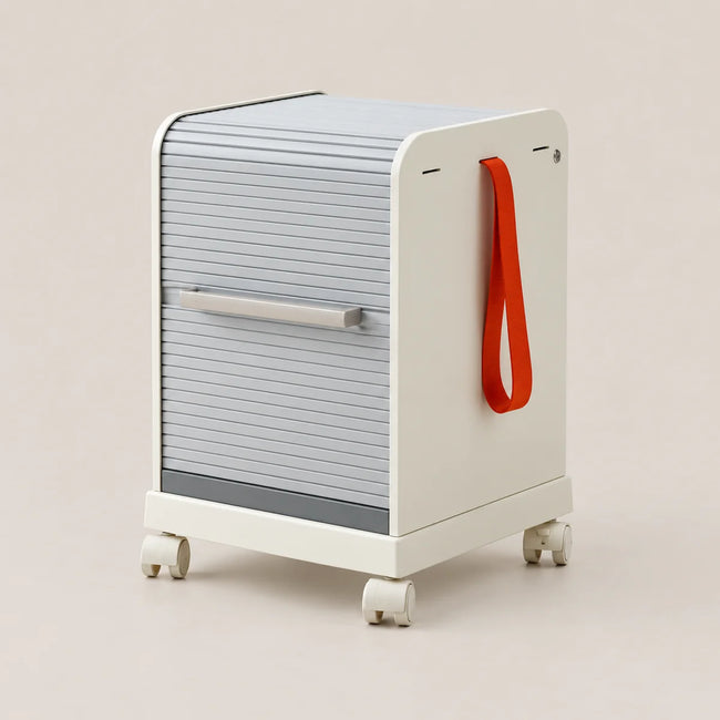 A modern rolling storage cabinet with a ribbed gray front panel, cream-colored side and base, a silver handle, and a bright red hanging loop, shown on a plain beige studio background.