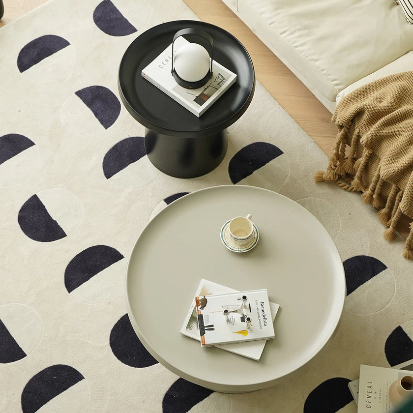 Round beige coffee table with a black side table on a patterned rug.