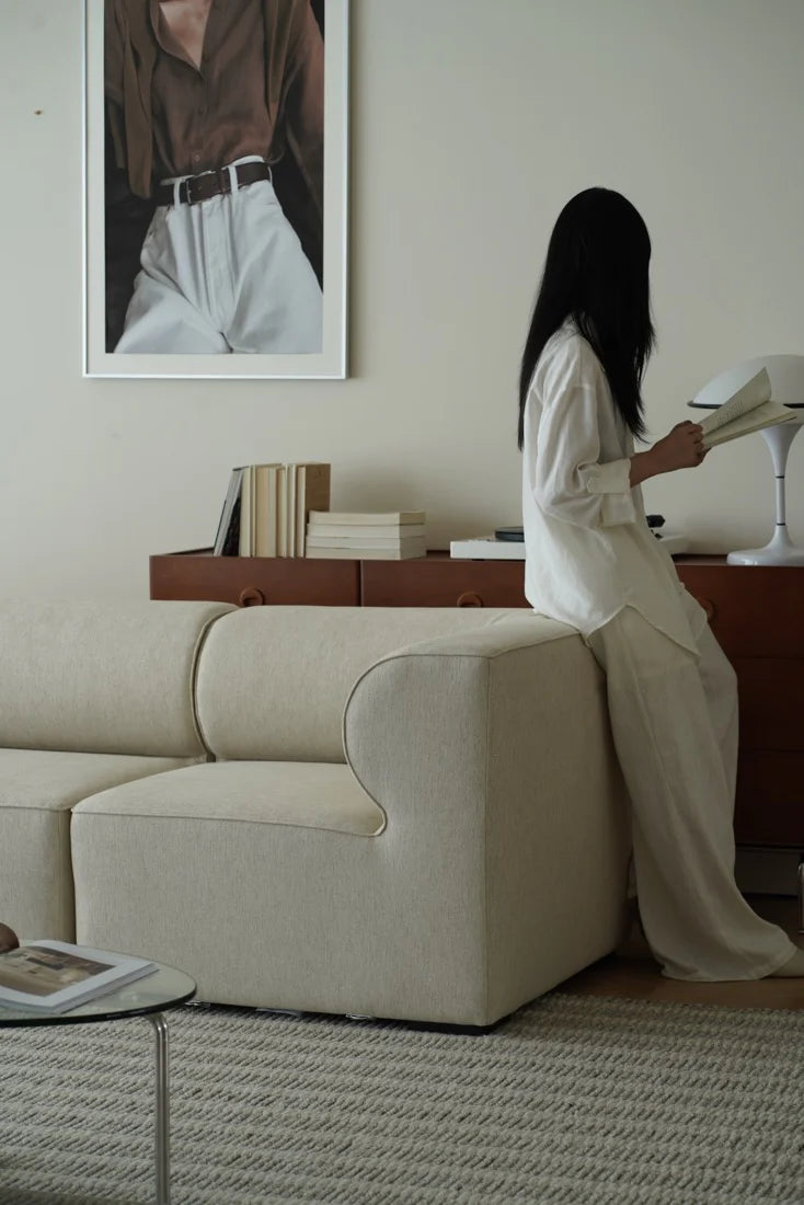 Woman sitting on a beige sofa reading a newspaper in a living room.