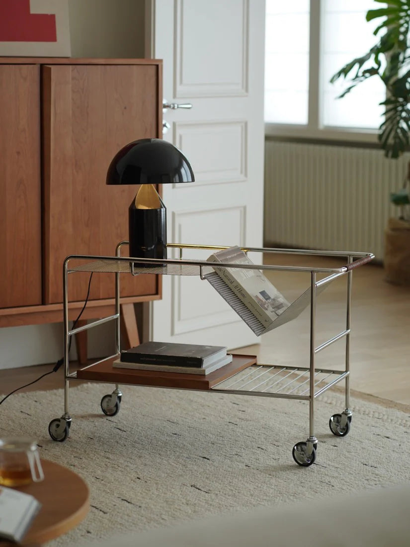 Modern interior with a metal trolley, black lamp, and books in a room with wooden cabinet and window.