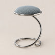 A modern stool with a textured light blue fabric cushion on a curved, polished chrome metal base, set against a solid beige background.