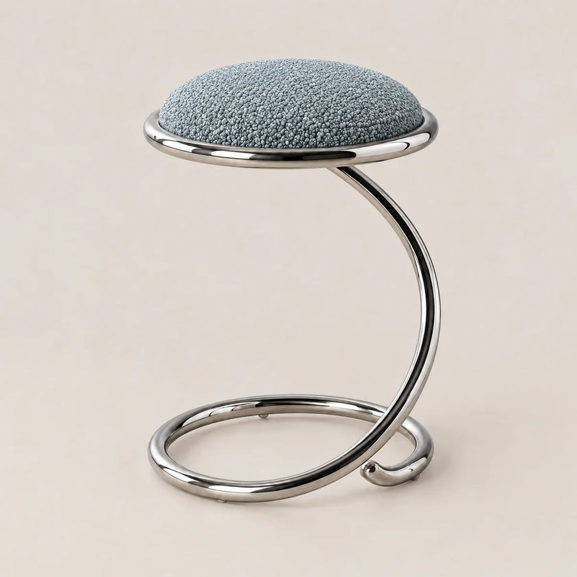 A modern stool with a textured light blue fabric cushion on a curved, polished chrome metal base, set against a solid beige background.