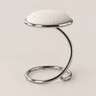 A modern stool with a textured off-white circular seat cushion and a curved, polished chrome metal base, set against a solid light beige background.
