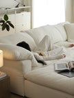 Person lying on a white couch under a blanket with an open book beside them.