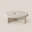 A minimalist, low-profile round ottoman or coffee table in a glossy off-white finish, featuring a central circular cutout on its top surface and a four-leg base, set against a solid, warm off-white background.