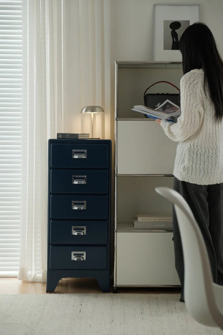 Person reading a book next to a blue filing cabinet in a room with white walls and a window.