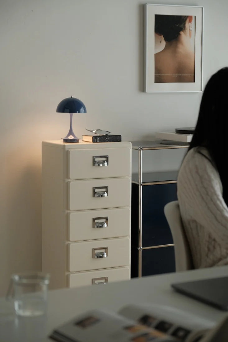 Filing cabinet with a lamp and books on top, framed picture on the wall, and a person sitting at a desk.