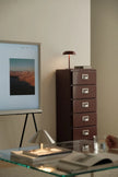 Modern interior with a glass table, brown filing cabinet, and framed artwork on a white wall.