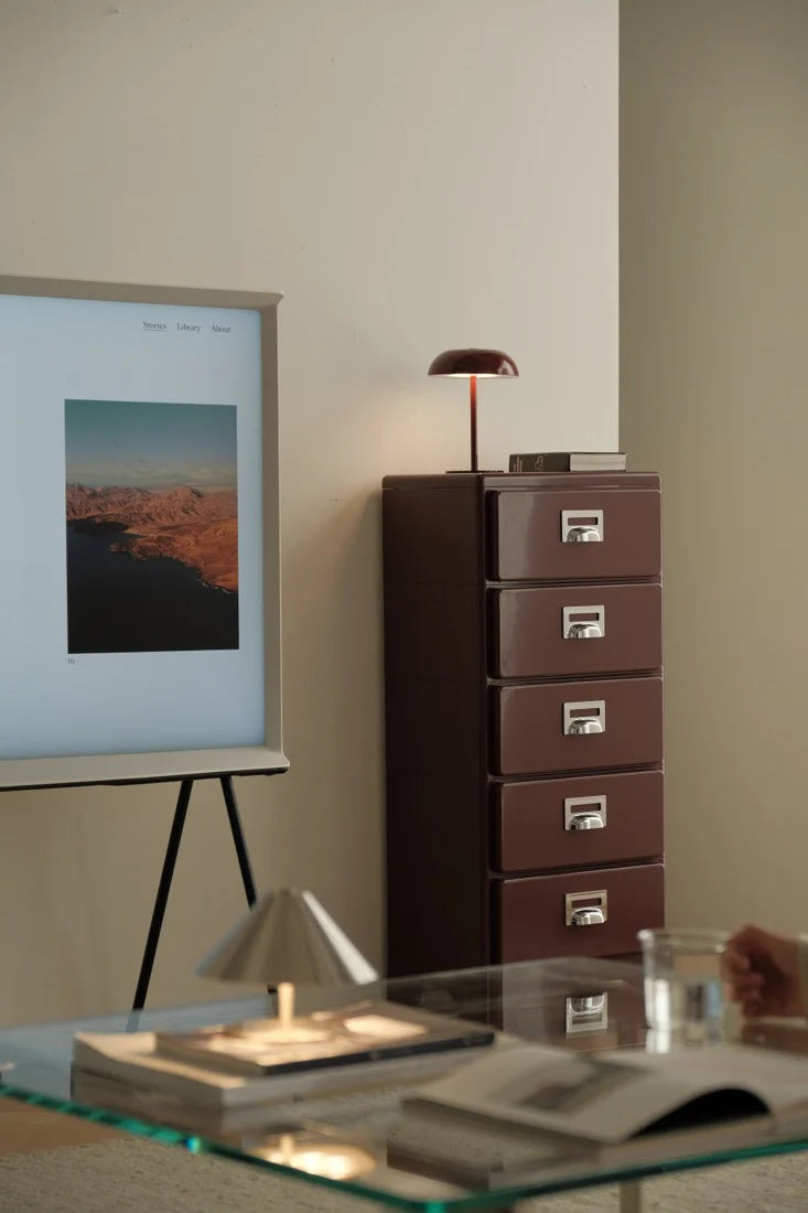 Modern interior with a glass table, brown filing cabinet, and framed artwork on a white wall.