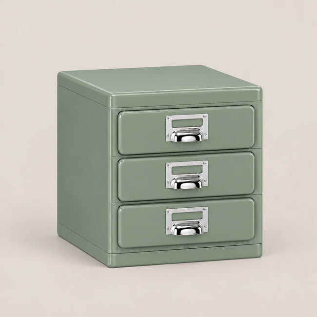 A three-drawer metal file cabinet in a muted sage green color, featuring silver metal pull handles with label slots, set against a solid light beige background.