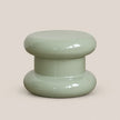 A glossy, mushroom-shaped pouf stool in a light sage green color, set against a solid, warm beige background.