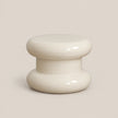 A glossy, two-tiered stool or small table in a warm cream or off-white color, set against a solid, neutral beige background.