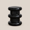A glossy black modern side stool with a stacked, tiered circular design, photographed against a solid light beige background.