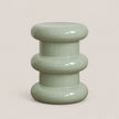 A glossy, modern three-tiered side stool in a light sage green color, set against a solid, warm off-white background.