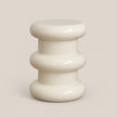 A glossy, modern side stool with a stacked, rounded organic design in a warm off-white/cream color, set against a solid light beige background.