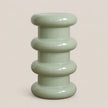 A glossy, ribbed ceramic stool in a pale sage green color, set against a solid, warm off-white background.