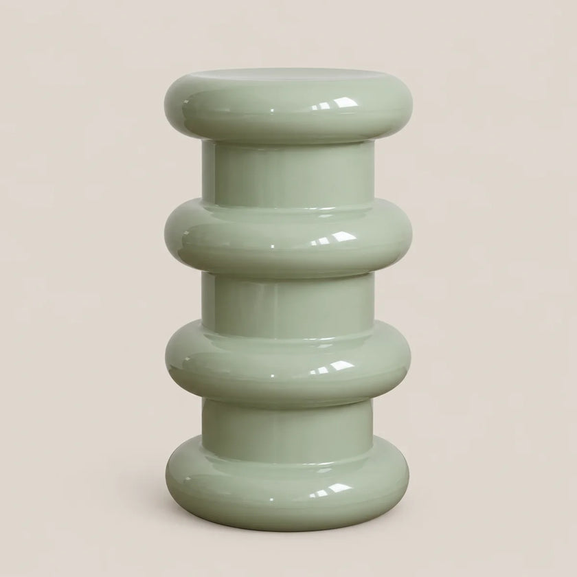 A glossy, ribbed ceramic stool in a pale sage green color, set against a solid, warm off-white background.