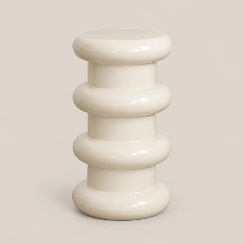 A glossy ceramic side stool with a stacked, ribbed design in a warm cream color, set against a solid light beige background.