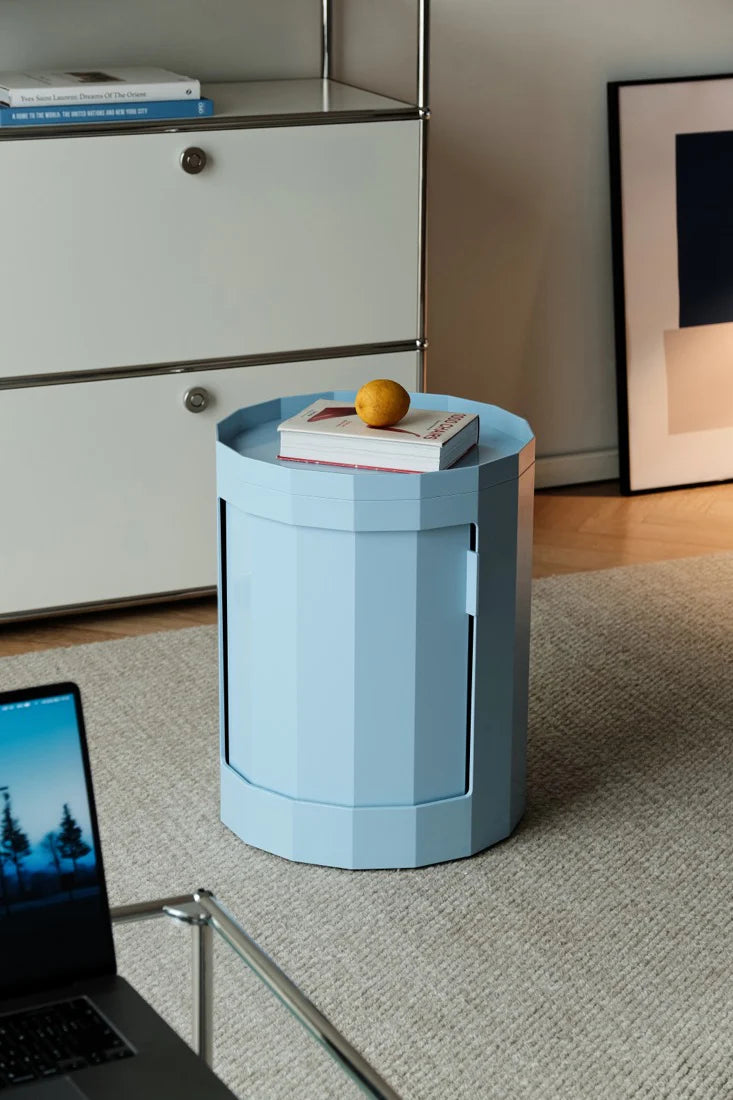 Blue cylindrical side table with a book and an orange on top, next to a laptop on a desk.