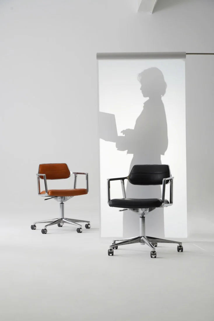Two office chairs, one black and one brown, in a minimalistic office setting with a person using a laptop in the background.