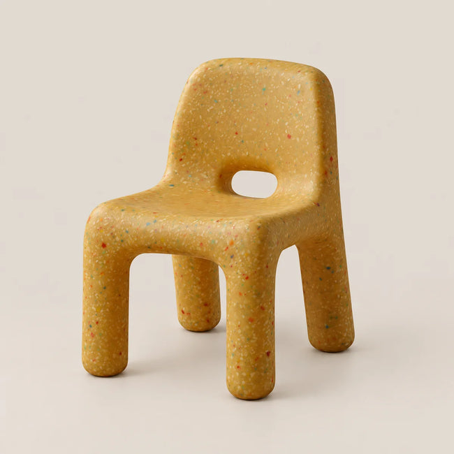 A small molded chair with a rounded backrest, armhole cutout, and four legs, shown on a plain warm off-white studio background. The chair has a mustard-yellow base with fine multicolored speckles throughout.