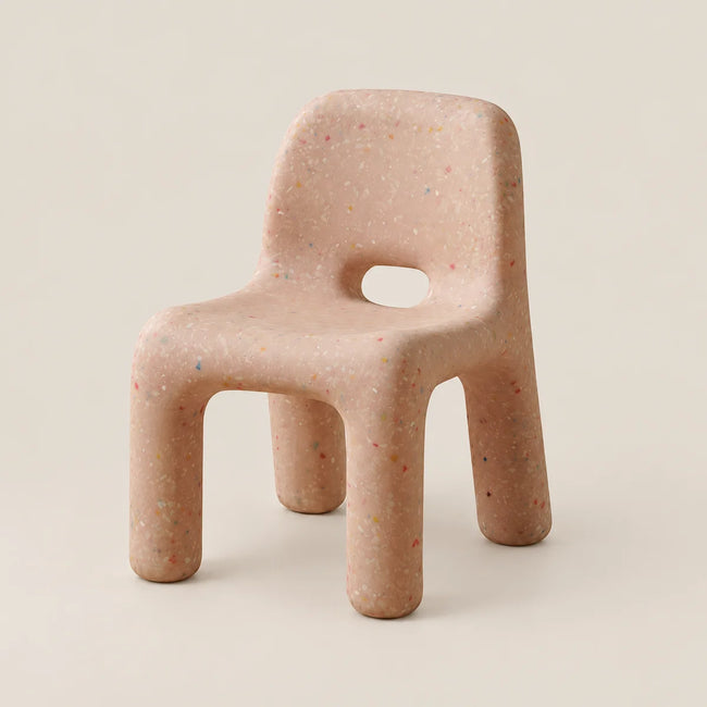 A small molded chair with a rounded backrest, thick legs, and a central cutout, shown on a plain warm off-white studio background. The chair has a light peachy-beige base with tiny multicolored flecks throughout.