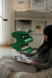 Woman reading a magazine on a couch next to a green side table with books and glasses.