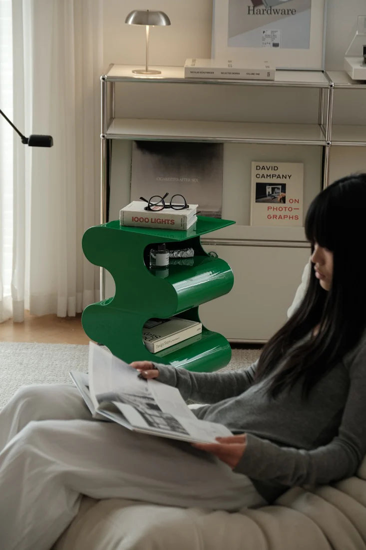 Woman reading a magazine on a couch next to a green side table with books and glasses.