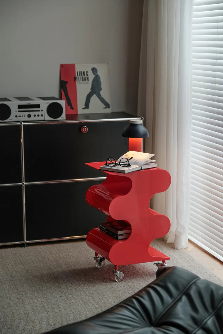 Red side table with books and glasses next to a black cabinet in a room with a window.