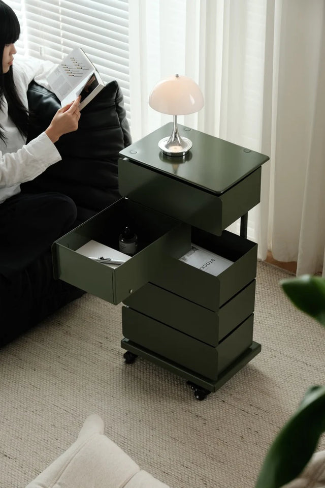 Person reading a book next to a black side table with open drawer in a room with curtains and a lamp.