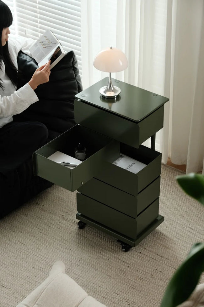 Person reading a book next to a black side table with open drawer in a room with curtains and a lamp.