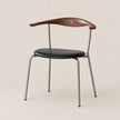 A mid-century modern dining chair featuring a curved dark wood backrest with a small gold cross in the center, a black upholstered seat, and a slim silver metal frame, set against a solid beige background.