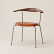 A mid-century modern dining chair featuring a curved dark wood backrest, a warm cognac leather seat cushion, and slim silver metal legs, set against a solid light beige background.