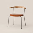 A mid-century modern dining chair featuring a curved natural wood backrest, a round cognac brown leather seat, and a slim silver metal frame, set against a solid warm beige background.