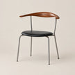 A mid-century modern dining chair featuring a curved wooden armrest, a black leather seat cushion, and a slim silver metal frame, set against a solid warm beige background.
