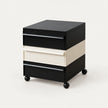 A modern three-drawer rolling storage cabinet with a black top and base, and a central cream-colored drawer front, shown on a light gray studio background.