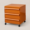 An orange plastic rolling file cabinet with three drawers and black caster wheels, photographed on a solid light beige background.