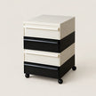 A four-drawer rolling storage cabinet with a modern color-blocked design. The cabinet features alternating cream and black drawer fronts and side panels, with a cream top surface and black caster wheels at the base, photographed against a solid light beige background.