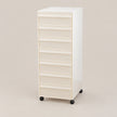 A tall, narrow white rolling storage cabinet with eight drawers and black caster wheels, photographed against a plain warm beige studio background.