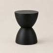 A minimalist black side table with a sculptural hourglass-like design, featuring a matte finish on a warm beige background.