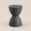 A minimalist hourglass-shaped side table in matte dark gray, featuring a circular top and a flared base, set against a solid light beige background.