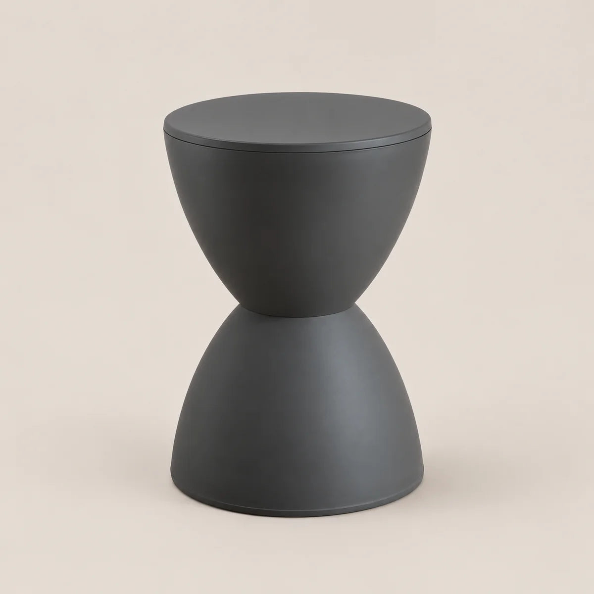 A minimalist hourglass-shaped side table in matte dark gray, featuring a circular top and a flared base, set against a solid light beige background.