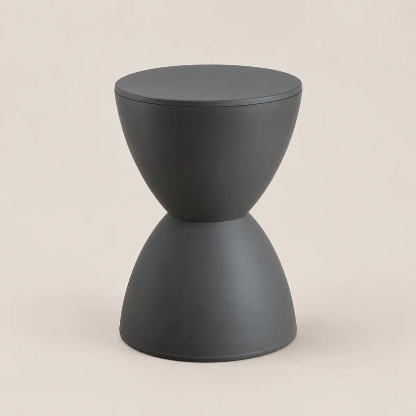 A minimalist hourglass-shaped side table in matte dark gray, featuring a circular top and a flared base, set against a solid light beige background.