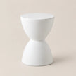 A minimalist white hourglass-shaped side table or stool with a smooth matte finish, set against a solid light beige background.