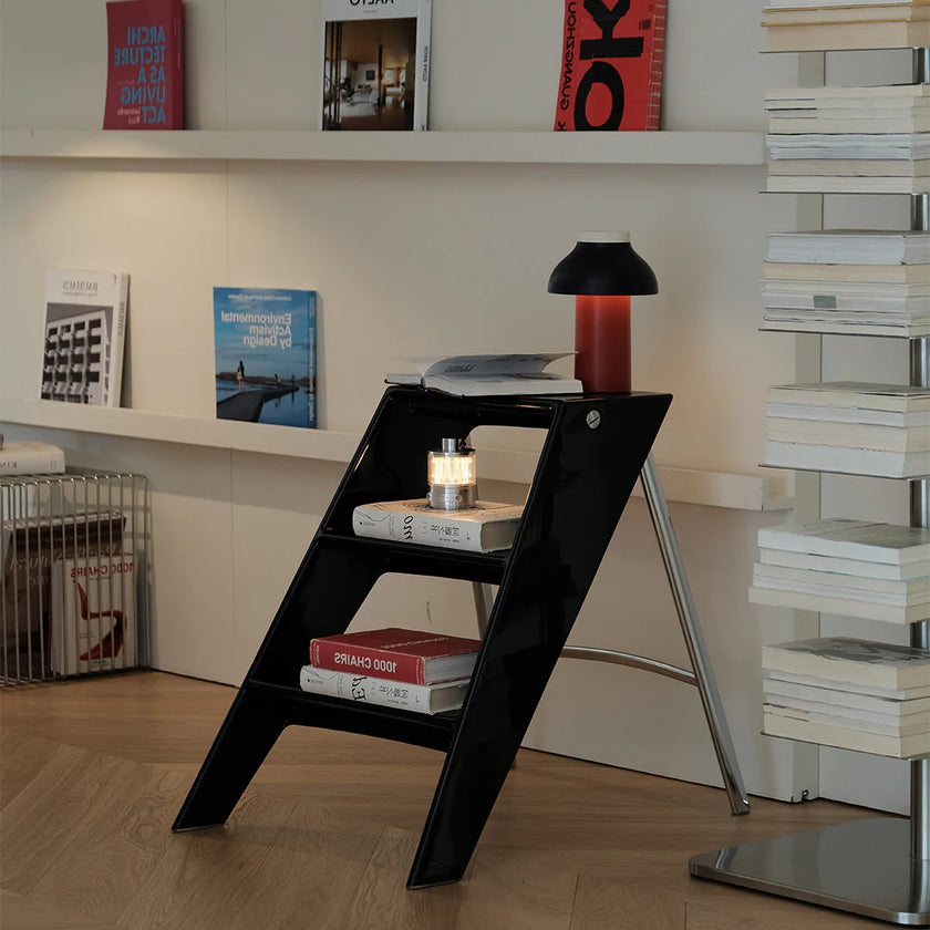 Black step ladder with books on a wooden floor, next to a white shelf with books and a lamp.