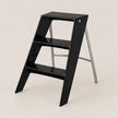 A modern black step stool with a glossy finish and silver metal support legs, shown on a clean, solid light beige background.