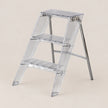 A three-step transparent acrylic step ladder with a silver metal support frame, shown on a solid light beige background.