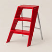 A bright red three-step ladder stool with a glossy finish and chrome metal legs, shown on a solid light beige background.