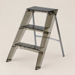 A three-step folding step ladder made of translucent smoky gray acrylic with black textured non-slip steps, shown on a solid light beige background.