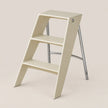 A minimalist two-step ladder stool in a warm cream color with a polished silver metal frame, shown on a solid light beige background.